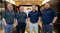Four smiling men in branded shirts standing in a hallway with elevators on both sides.