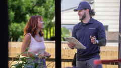 A woman smiles and speaks with a clipboard-holding worker in a cap outside near a wooden fence.