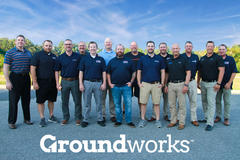 Group of people standing outdoors wearing Groundworks shirts, with a clear sky and trees in the background.