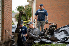 Three construction workers in blue shirts fixing a foundation near brick buildings, surrounded by equipment and tools.