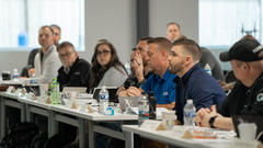 People sitting at a conference table, attentively listening to a speaker, with notepads and drinks on the table.