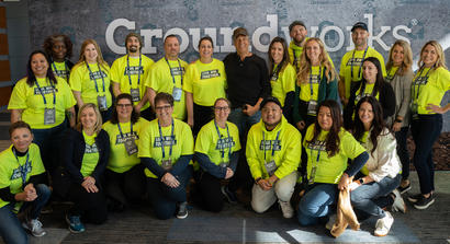 A group of people in bright shirts pose in front of a "Groundworks" banner indoors.