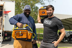 Two smiling men, each holding an orange tool bag. A truck and building are in the background.