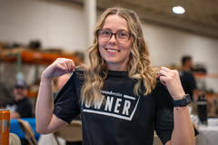 Smiling person with glasses holding a "Groundworks Owner" shirt in a room with tables and chairs.