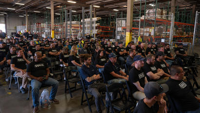 A crowd of people wearing matching black shirts sit on chairs in a warehouse setting.