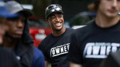A man in a cap and sunglasses smiles, wearing a shirt that says "Owner.