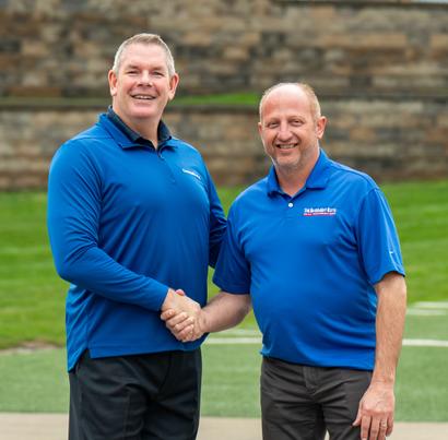 Two men in blue shirts shaking hands and smiling outdoors with a stone wall and grass in the background.