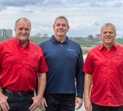 Three men stand outdoors, two in red shirts and one in blue, with a cityscape in the background.