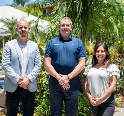 Three people standing outdoors in front of palm trees, smiling at the camera.