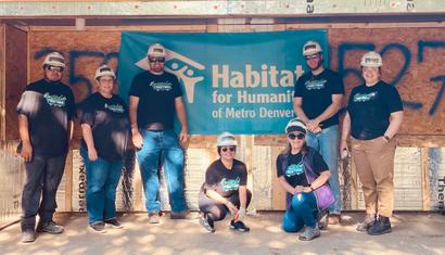 A group of volunteers wearing hard hats poses in front of a Habitat for Humanity sign.