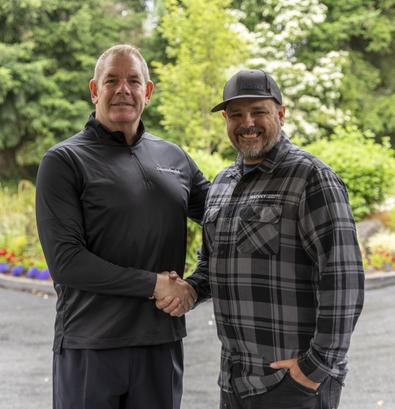 Two men smiling and shaking hands outside, surrounded by greenery and flowers.