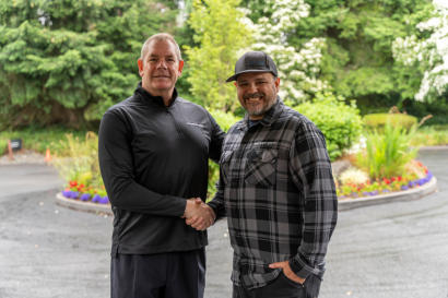 Two men shake hands outside on a paved road with trees and colorful flowers in the background.