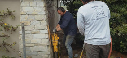 Two men using construction equipment beside a stone wall. One wears headphones; the other has “Rockbuilt Rocks!” on his shirt.