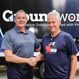 Two men smiling and shaking hands in front of a "Groundworks" sign.
