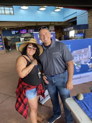 Two people posing and smiling at an outdoor event, standing near a table with brochures and a display banner.