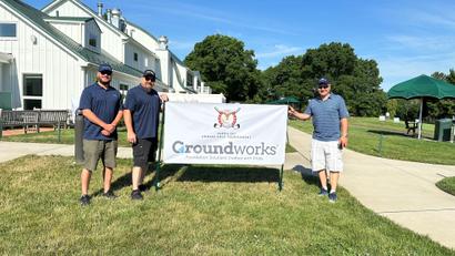Three people stand beside a Groundworks banner at a golf tournament on a sunny day.