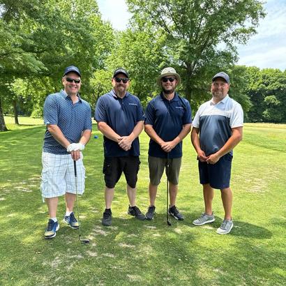 Four men standing on a golf course wearing casual golf attire, holding clubs, with trees in the background.