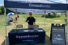 Person in sunglasses stands at a Groundworks booth outdoors, under a tent on a sunny day.