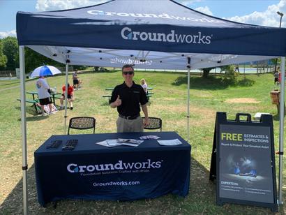 Person in sunglasses stands at a Groundworks booth outdoors, under a tent on a sunny day.