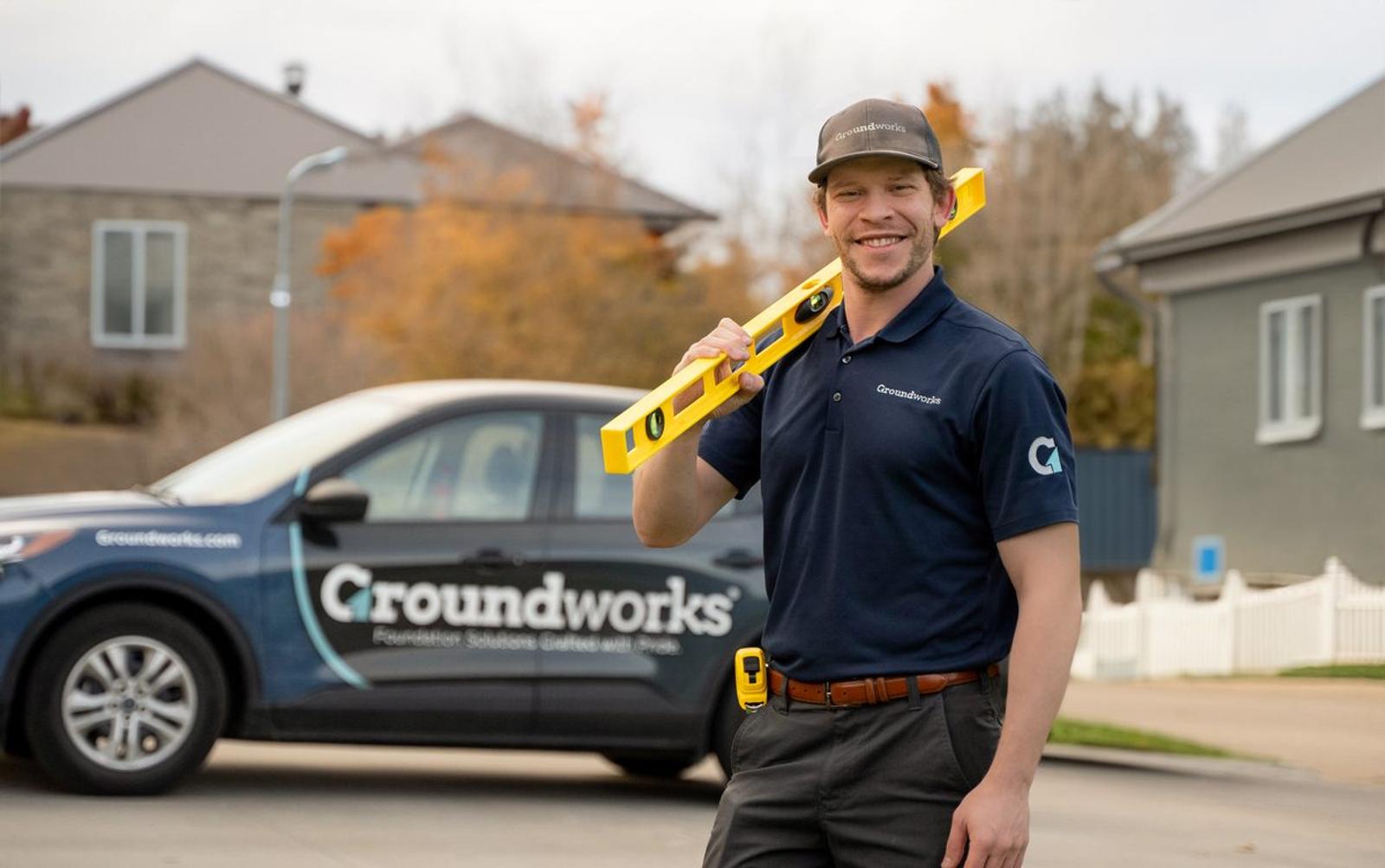 Smiling worker in Groundworks uniform holding a level, standing in front of a company vehicle and houses.