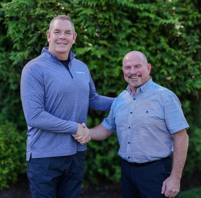 Two men shaking hands and smiling in front of green foliage.