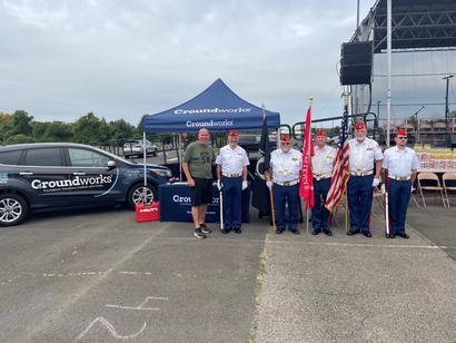 A group of six people in uniform and one in casual attire stand in front of a Groundworks tent and vehicle.