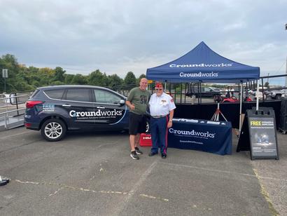 Two people stand smiling in front of a Groundworks booth and branded vehicle at an outdoor event.