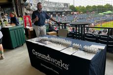 Man stands by a Groundworks table with flyers and products at a baseball stadium, giving a thumbs-up.