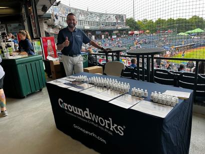 Man stands by a Groundworks table with flyers and products at a baseball stadium, giving a thumbs-up.