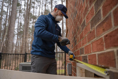 Person measuring with a tape measure against a brick wall, wearing a mask and gloves, with trees in the background.