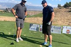 Two men standing on a golf course with clubs, smiling, signs in the background.