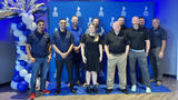 A group of people posing in front of a BBB backdrop with blue and white balloons.