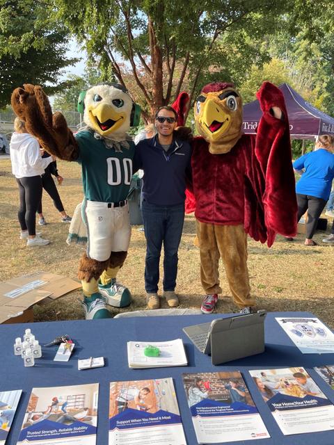 A person poses with two bird mascots at an outdoor event, surrounded by informational pamphlets on a table.