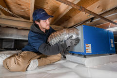Technician installing ductwork under a floor, wearing gloves and a cap in a wood-beamed space.