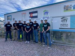 A group of people in helmets and t-shirts stand in front of a Habitat for Humanity trailer.