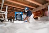 Worker in protective gear sets up equipment in a crawl space with plastic sheeting on the floor.