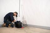 A worker kneels on the floor installing equipment near a wall outlet and white pipes in a basement.
