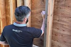 Man writing on a wooden wall frame, wearing a black "Goodwerks" shirt.