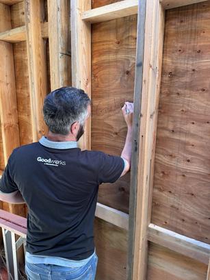 Man writing on a wooden wall frame, wearing a black "Goodwerks" shirt.