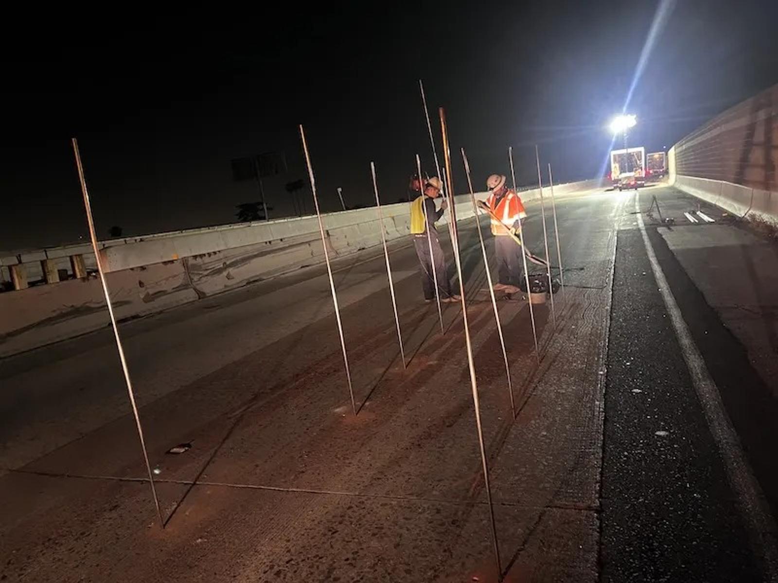 Workers in safety gear set up vertical rods for roadway stabilization on a closed highway at night, with a truck in the background.