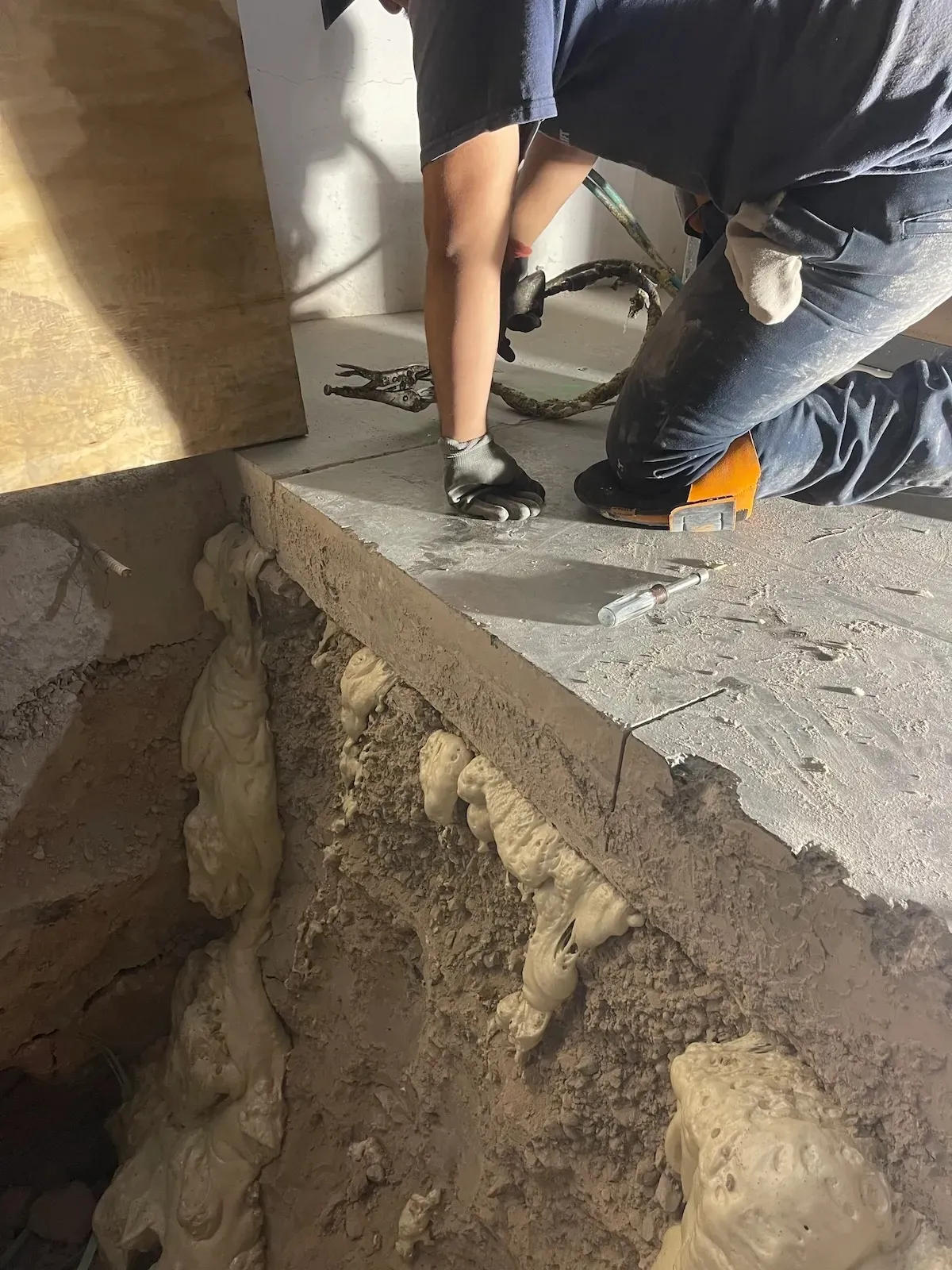 A person kneels on concrete by a large hole with exposed foam material, wearing gloves and holding a tool for void remediation.