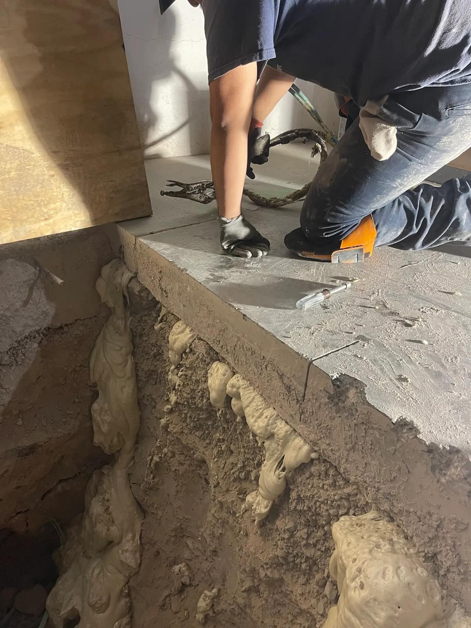 A person kneels on concrete by a large hole with exposed foam material, wearing gloves and holding a tool for void remediation.