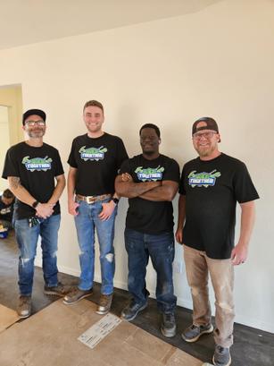Four men in matching shirts stand in a room under renovation, smiling at the camera.