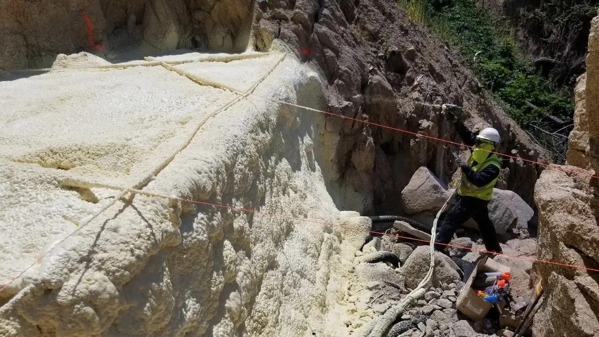 A worker in safety gear sprays foam for structural void filling on a rock surface in a rocky outdoor setting.