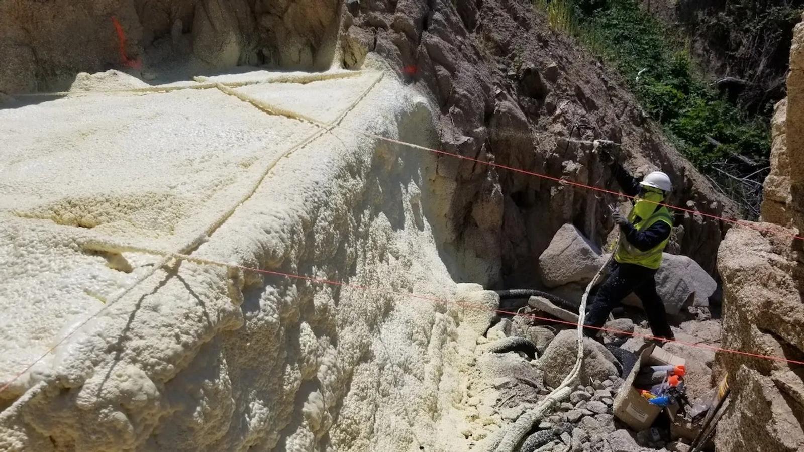 A worker in safety gear sprays foam for structural void filling on a rock surface in a rocky outdoor setting.