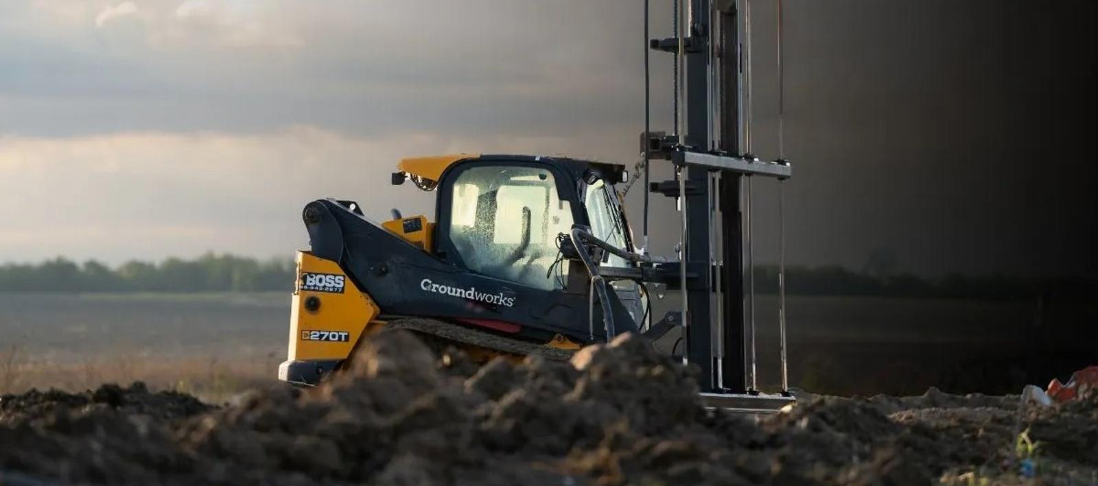 A construction machine operates on a muddy field with a cloudy sky in the background.