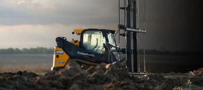 A construction machine operates on a muddy field with a cloudy sky in the background.