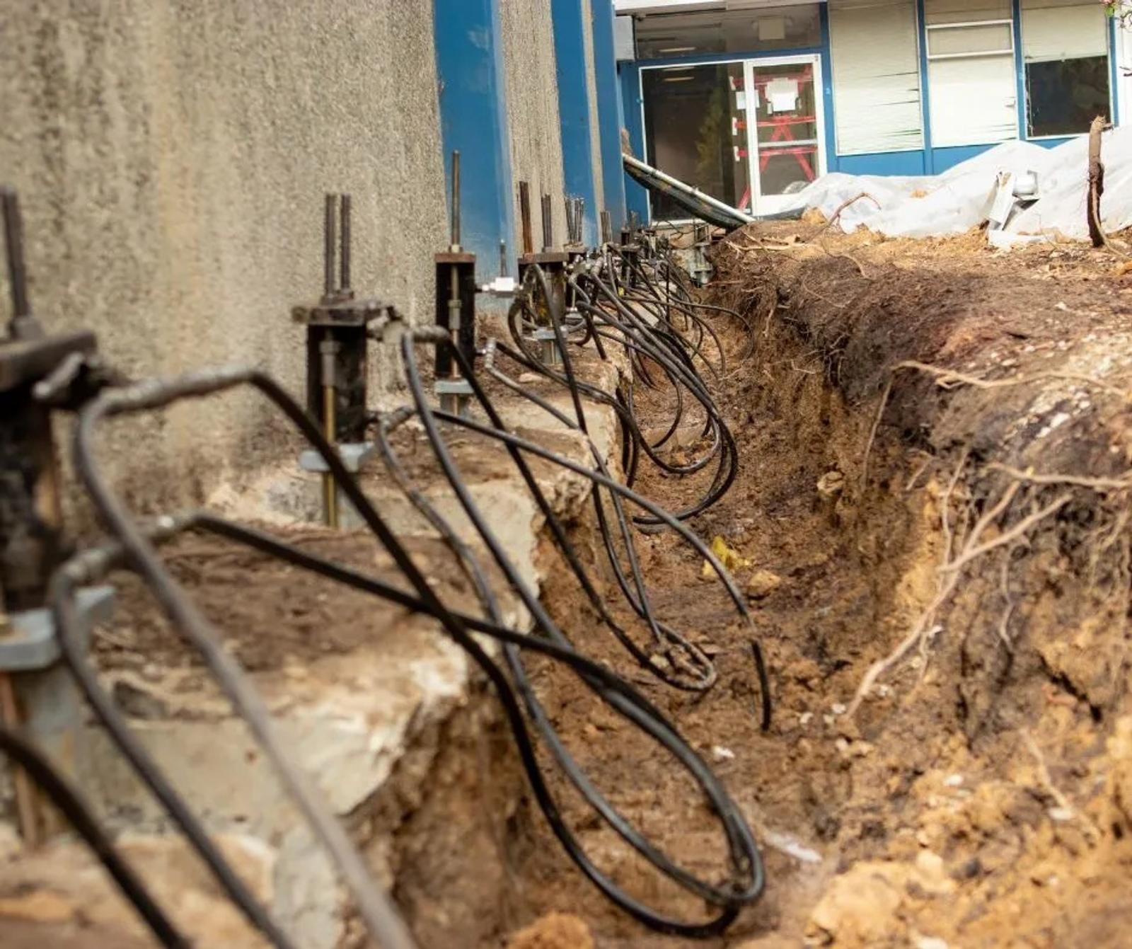 Close-up of a trench with exposed cables and grounding rods next to a building's commercial foundation underpinning.