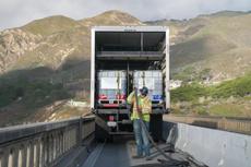 A worker in safety gear unloads tanks from a truck on a scenic mountain road bridge, showcasing Infrastructure Rehabilitation Services by Groundworks.