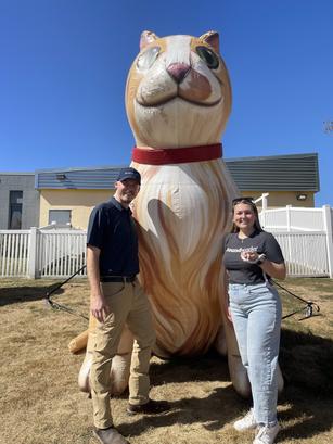 Two people stand smiling next to a large inflatable cat with a red collar outdoors.